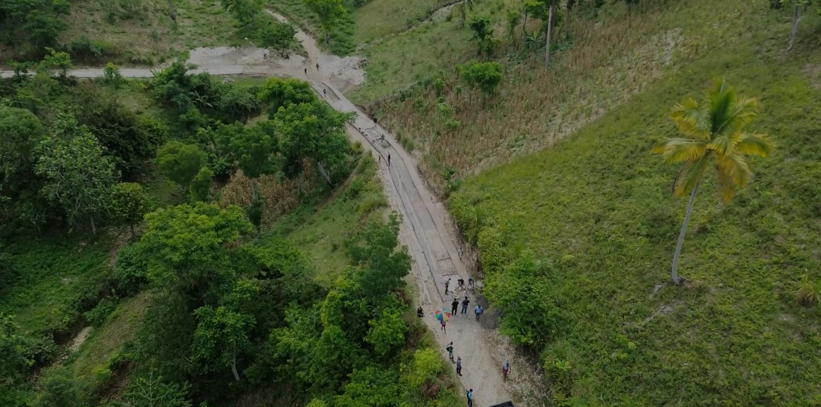 An aerial view of a dirt road winding through a green, hilly landscape, with several people walking along the road and dense vegetation on both sides.