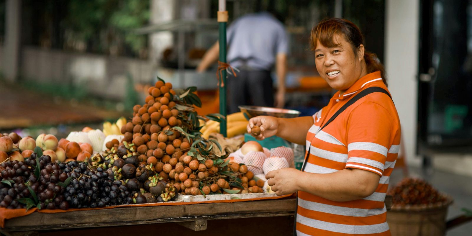 Woman in an orange and white striped shirt selecting fresh fruits at an outdoor market stall filled with lychees, grapes, peaches, and assorted produce.