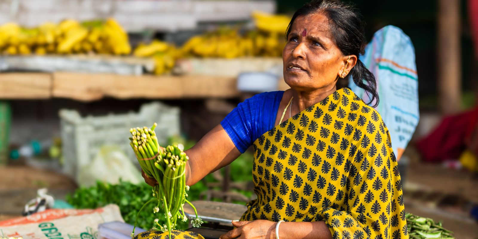 Woman in a yellow and blue patterned garment holding green vegetables at a market stall with bananas and assorted produce in the background.