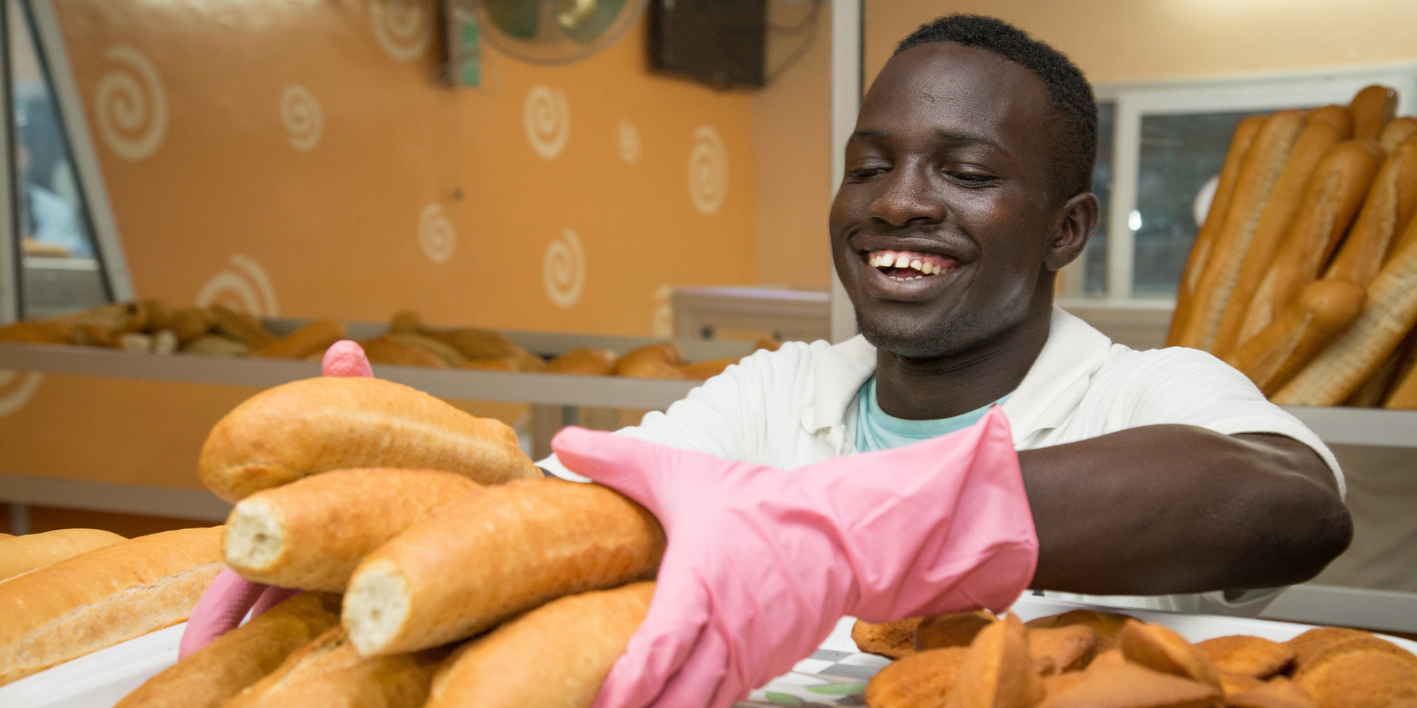 Man wearing pink gloves arranging fresh loaves of bread in a bakery with decorative swirl-patterned walls and shelves filled with assorted baked goods.