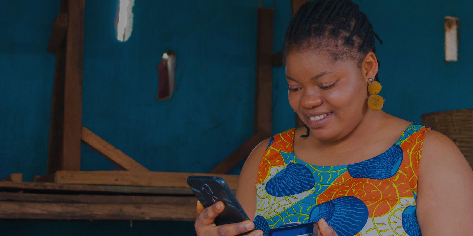 Woman in a colorful patterned dress with braided hair holding a smartphone indoors, against a backdrop of wooden structures and blue walls.