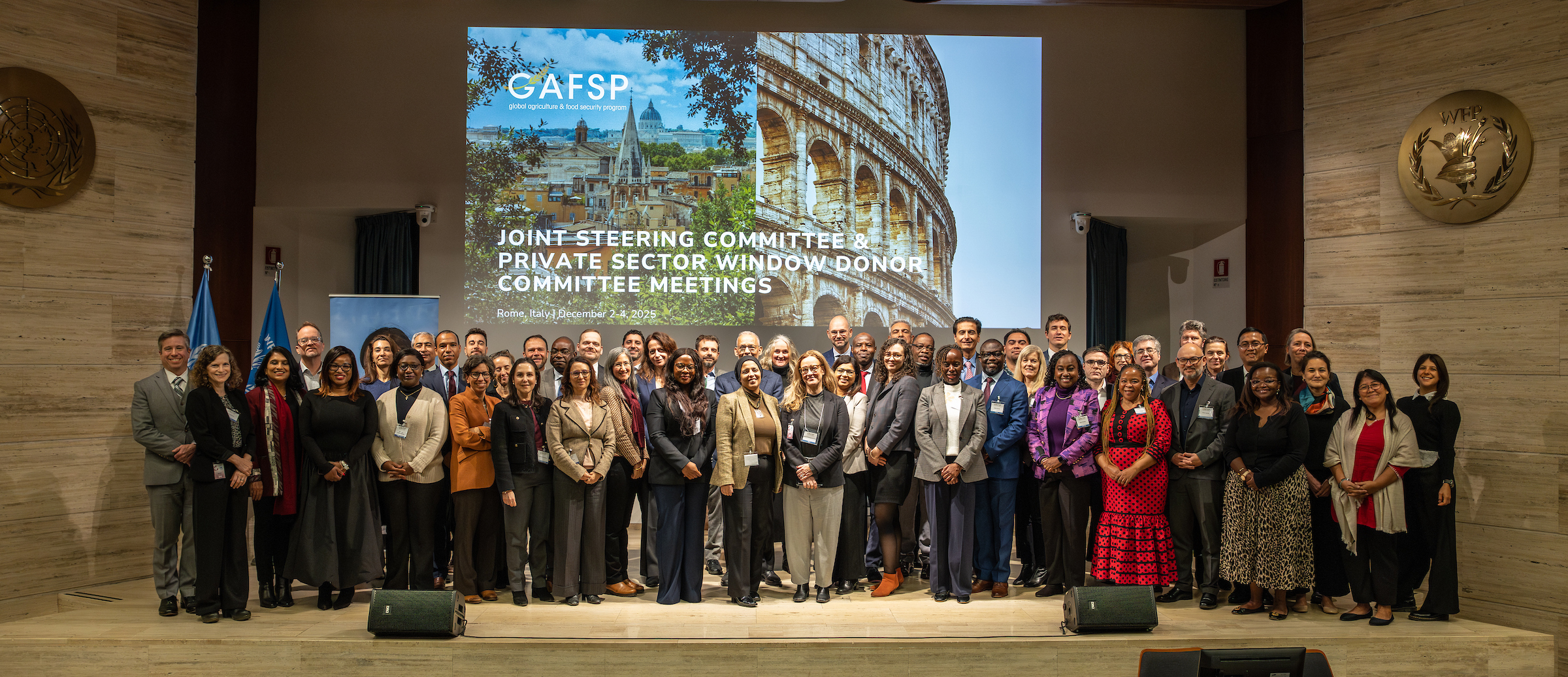 A large group of people standing on a stage in a conference room, posing for a photo in front of a large screen. The screen displays the GAFSP logo and text that reads: “Joint Steering Committee & Private Sector Window Donor Committee Meetings, Rome, Italy, December 2023.” The background image on the screen shows landmarks including the Colosseum and a cityscape. Two UN-style emblems are mounted on the walls, and flags are visible on the left side.