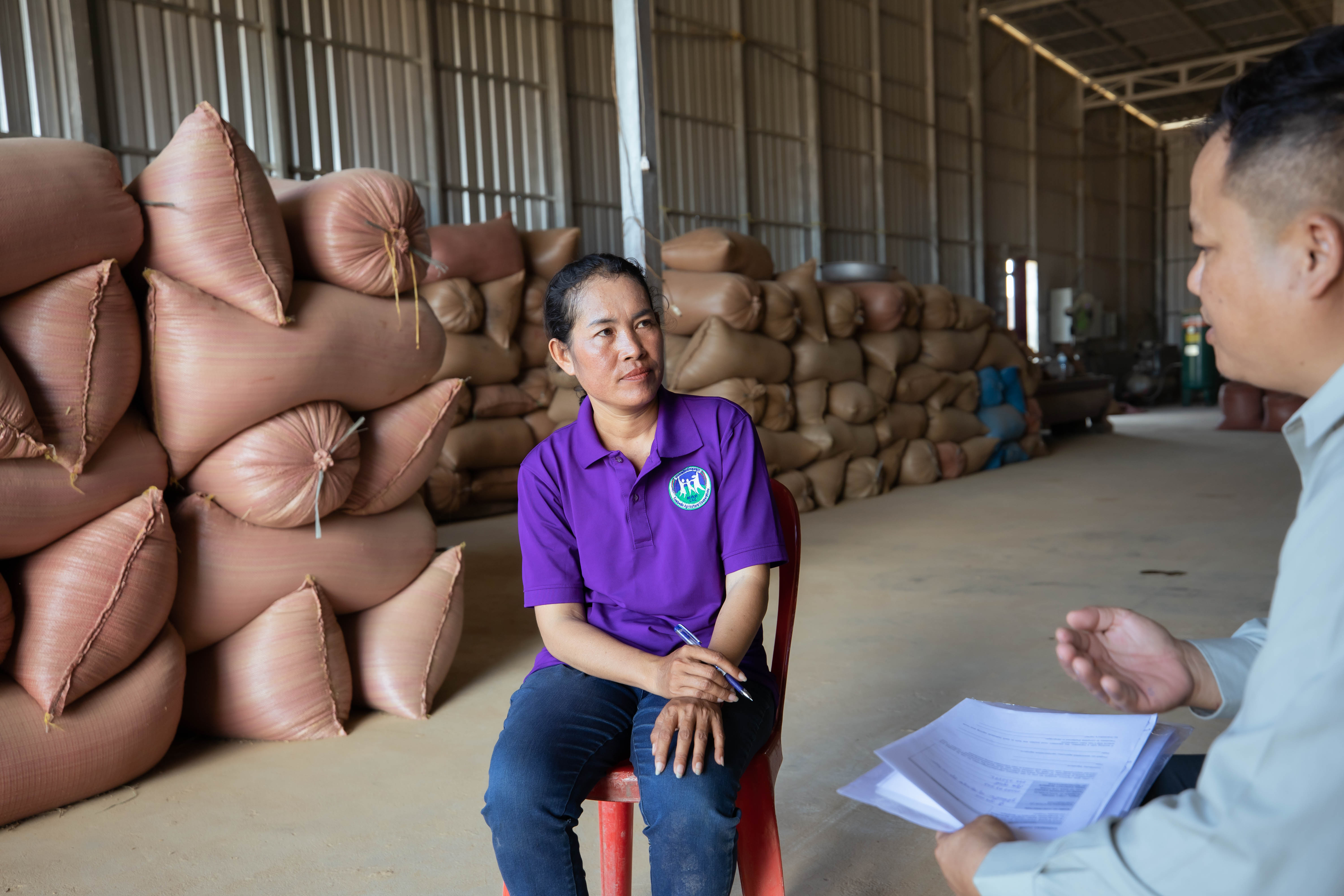 A person sitting on a red stool inside a large warehouse speaks with another person holding papers. Stacks of filled sacks are piled high in the background along the walls of the storage space.