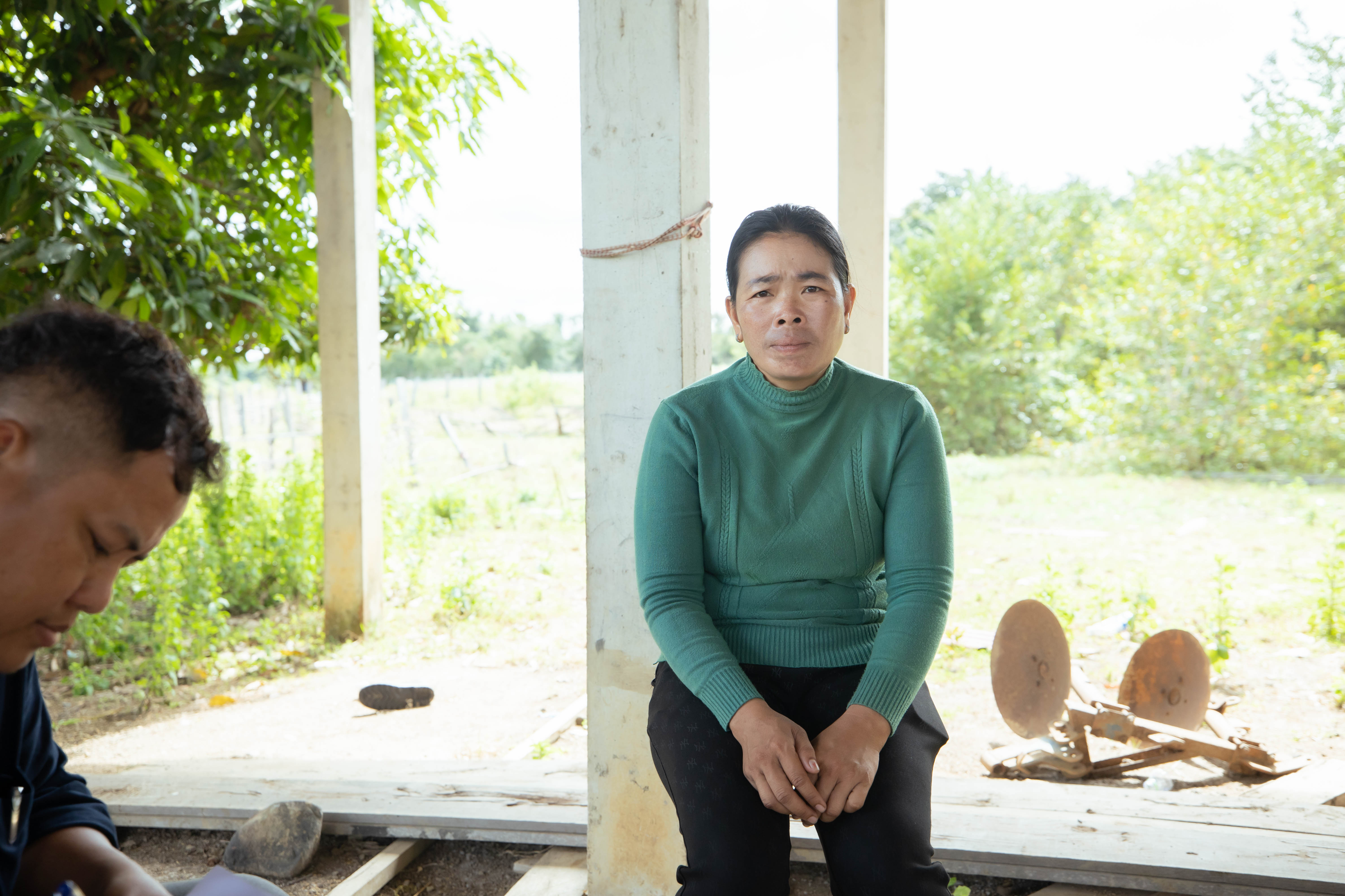 A person wearing a green sweater sits on a concrete porch supported by pillars, facing the camera. Behind them is a sunlit rural landscape with trees and open ground. Another person is partially visible in the foreground on the left, looking downward, while farming tools or metal equipment lie on the ground to the right.