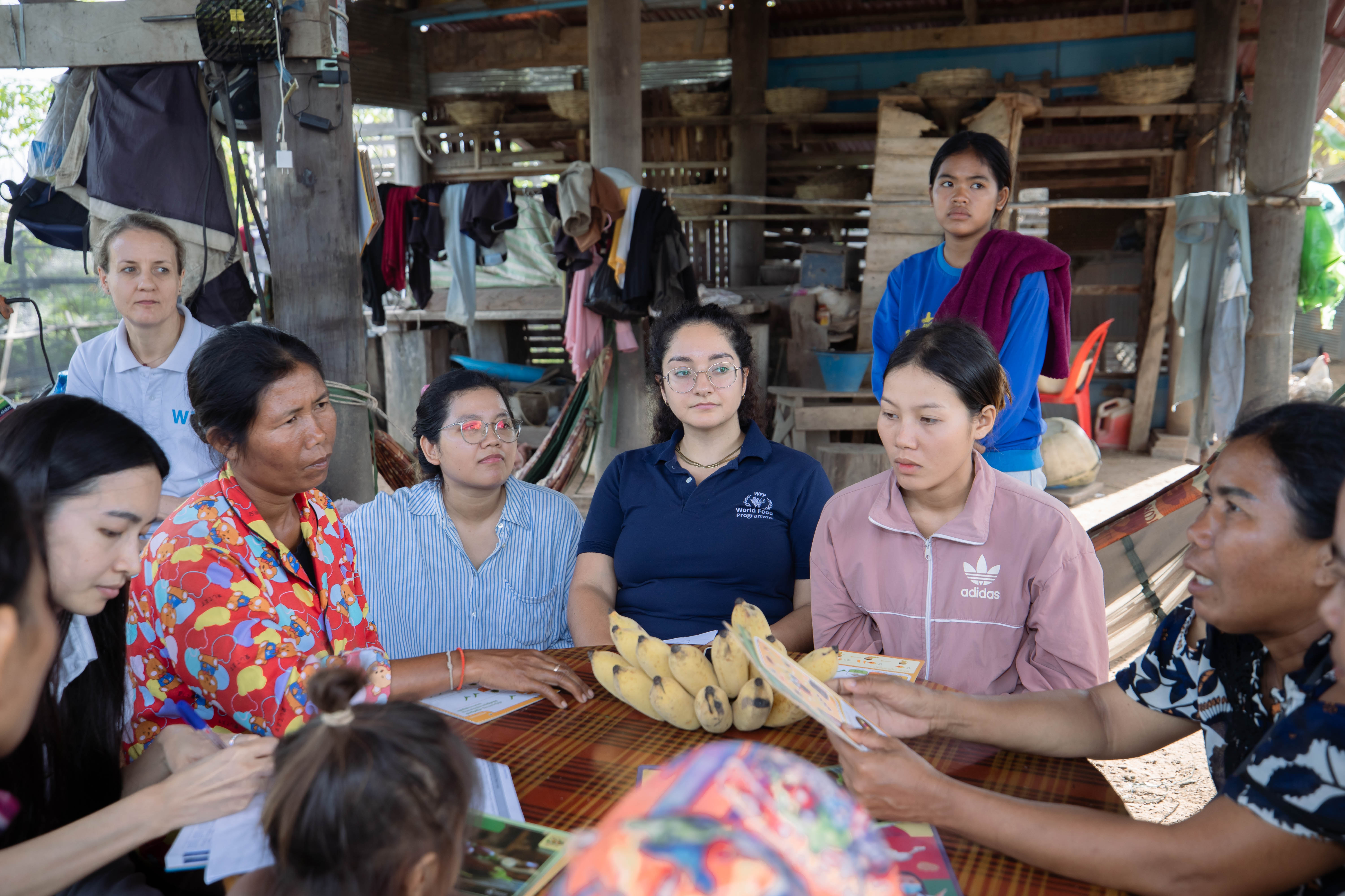A group of people sit around a table inside a simple open-sided home...