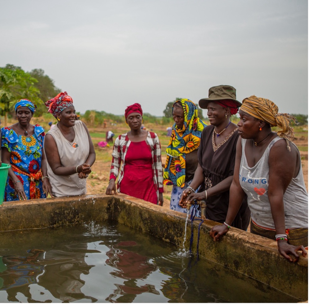 A group of people stand gathered around a rectangular outdoor water basin. They are wearing colorful, patterned clothing and head coverings, and appear to be engaged in conversation or observing the water. The setting is an open, rural area with soil, scattered vegetation, and trees visible in the background under an overcast sky.