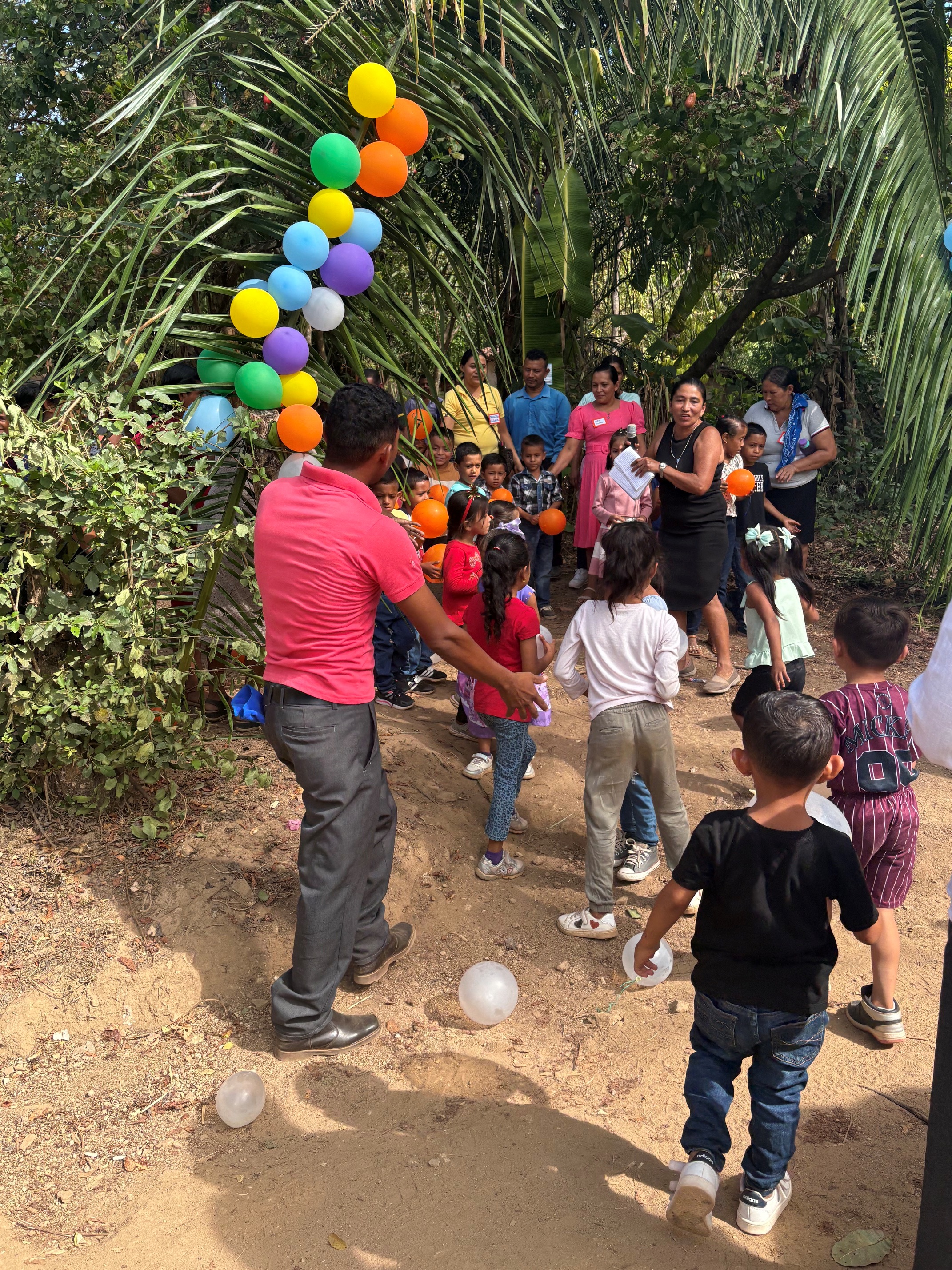 A group of children and adults gather along a dirt pathway in a tropical outdoor setting surrounded by palm trees and dense greenery. Colorful balloons are attached to an arch made from palm fronds, creating a festive entrance. Several children hold orange balloons while adults stand nearby. The scene appears to be a community celebration or activity taking