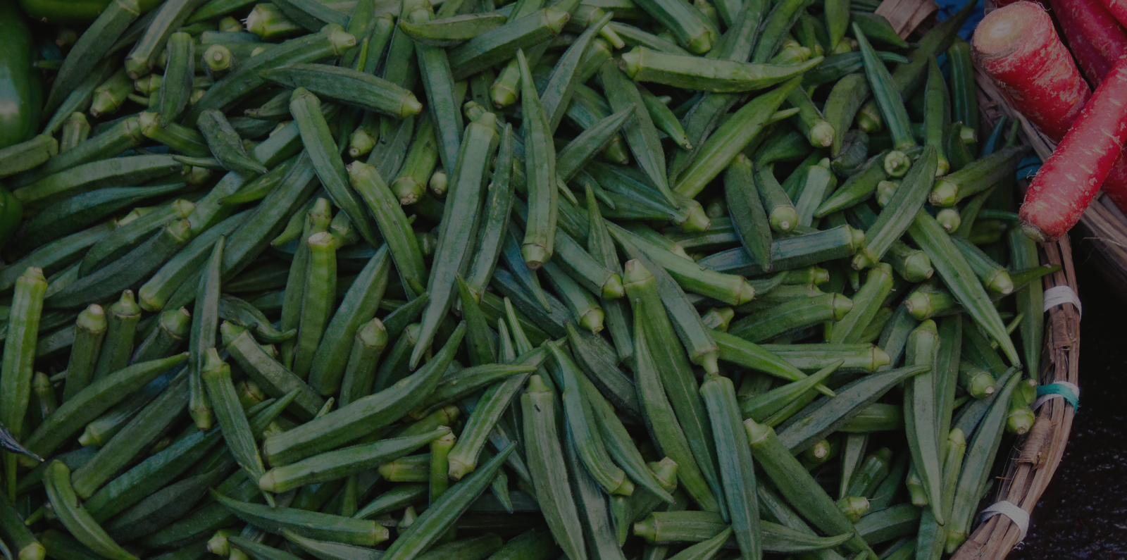 A basket filled with fresh green okra pods, elongated and slightly ridged, with a few red carrots peeking in from the right side, adding a pop of contrasting color.
