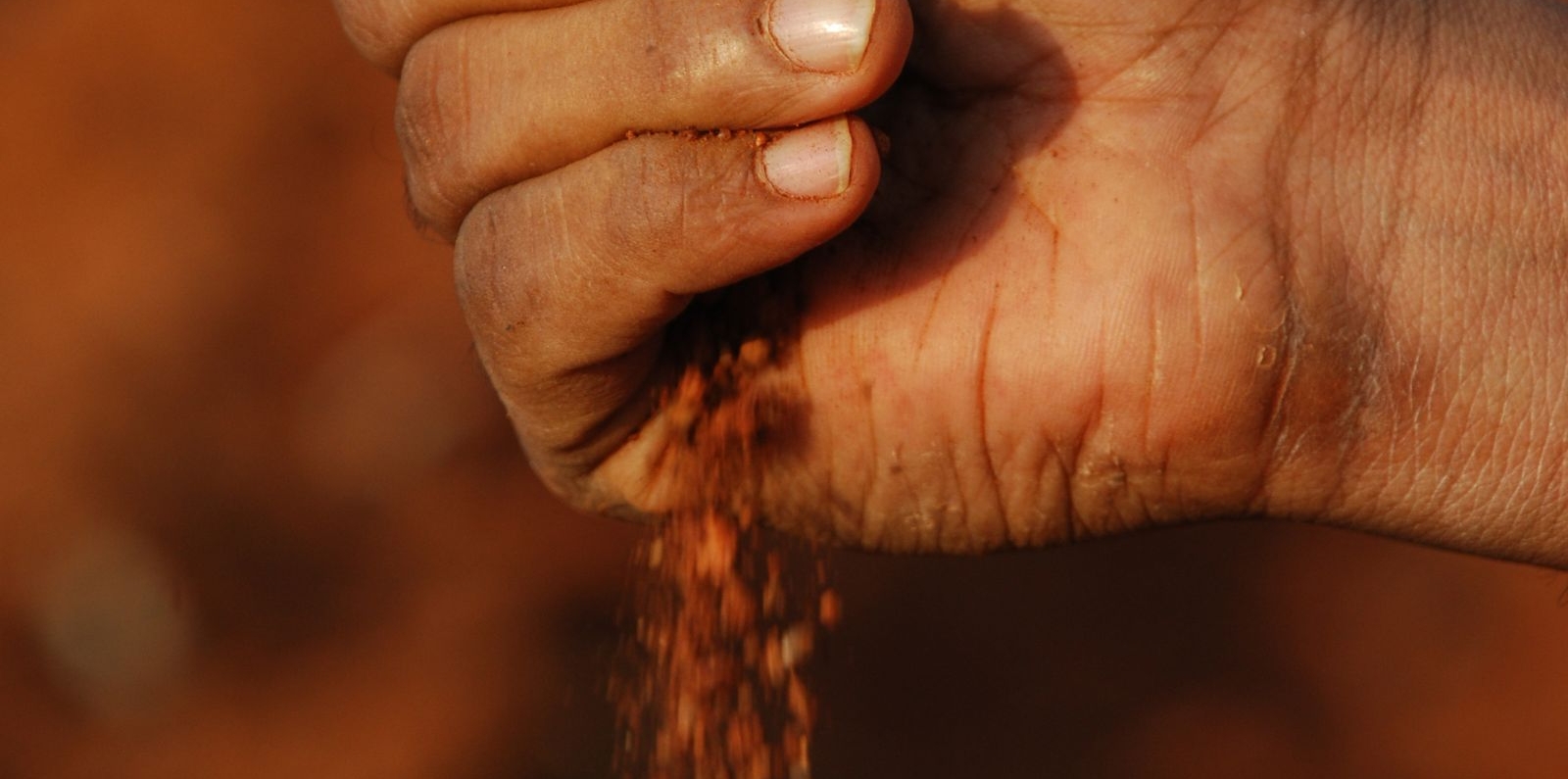 A close-up of a hand holding reddish-brown soil, letting it fall through the fingers against a blurred earthy background.