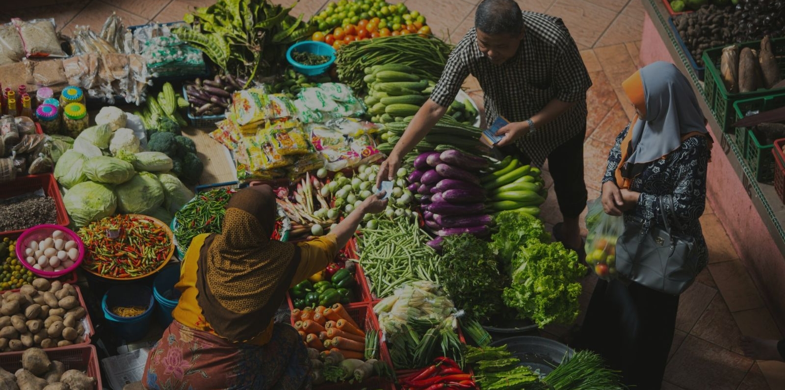 Three people engaged in a transaction at a vibrant market stall filled with fresh vegetables like cabbages, eggplants, green beans, chilies, and lettuce, along with packaged goods.