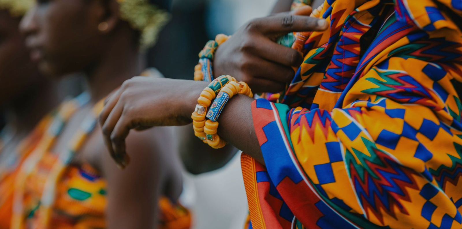 Close-up of a person wearing a brightly colored Ghanian traditional patterned garment and orange beaded bracelets, with another person in similar attire visible in the background.