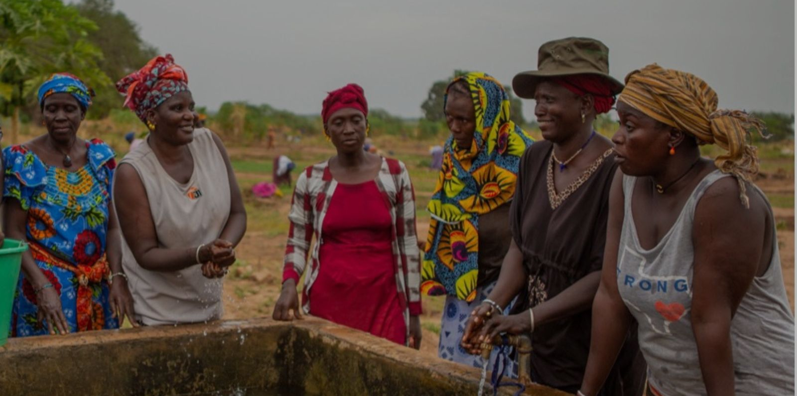 A group of people stand gathered around a rectangular outdoor water basin. They are wearing colorful, patterned clothing and head coverings, and appear to be engaged in conversation or observing the water. The setting is an open, rural area with soil, scattered vegetation, and trees visible in the background under an overcast sky.
