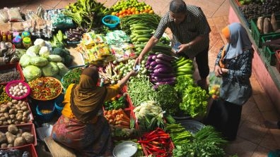 Three people engaged in a transaction at a vibrant market stall filled with fresh vegetables like cabbages, eggplants, green beans, chilies, and lettuce, along with packaged goods.