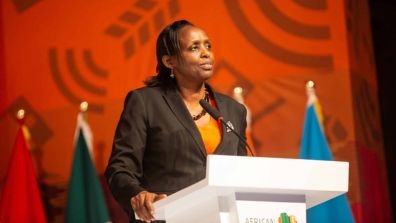 Agnes Kalibata standing at a podium delivering a speech at a formal event, wearing a dark suit and orange top. The podium has a logo with green and orange elements. Several colorful flags are displayed in the background against an orange backdrop with abstract patterns.