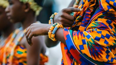 Close-up of a person wearing a brightly colored Ghanian traditional patterned garment and orange beaded bracelets, with another person in similar attire visible in the background.