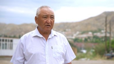 A person standing outdoors in front of a mountainous landscape, wearing a white short-sleeved shirt. The background shows hills, scattered buildings, and a partly cloudy sky.