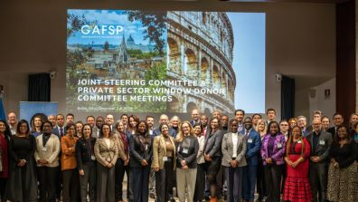 A large group of people standing on a stage in a conference room, posing for a photo in front of a large screen. The screen displays the GAFSP logo and text that reads: “Joint Steering Committee & Private Sector Window Donor Committee Meetings, Rome, Italy, December 2023.” The background image on the screen shows landmarks including the Colosseum and a cityscape. Two UN-style emblems are mounted on the walls, and flags are visible on the left side.