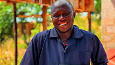 A person wearing a dark blue work uniform stands outdoors in a sunlit area with trees and wooden structures in the background.