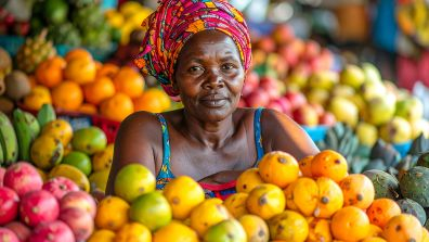 A market vendor wearing a colorful patterned headwrap sits behind a large display of fresh fruits, including mangoes, papayas, and other vibrant produce at an outdoor market stall.
