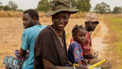 A group of people sit together outdoors on a dry, grassy field. They are seated in a row, some holding tools, including a yellow-handled item in the foreground. They wear a mix of hats, patterned clothing, and casual attire. Trees and tall grass are visible in the background under an overcast sky.