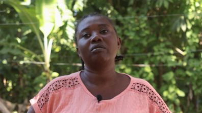 A person wearing a textured peach-colored top with embroidered details stands outdoors in a lush, green setting. The background includes leafy plants and soft natural light.