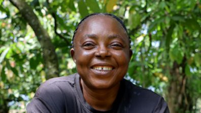 A farmer smiling directly at the camera outdoors, with green foliage in the background.