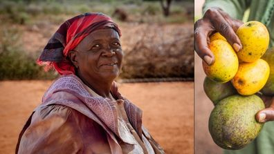 Close-up of a person wearing a red and purple patterned headscarf and layered clothing, seated outdoors on reddish-brown ground. The background is softly blurred, showing an arid landscape and another partially visible person at the edge of the frame.